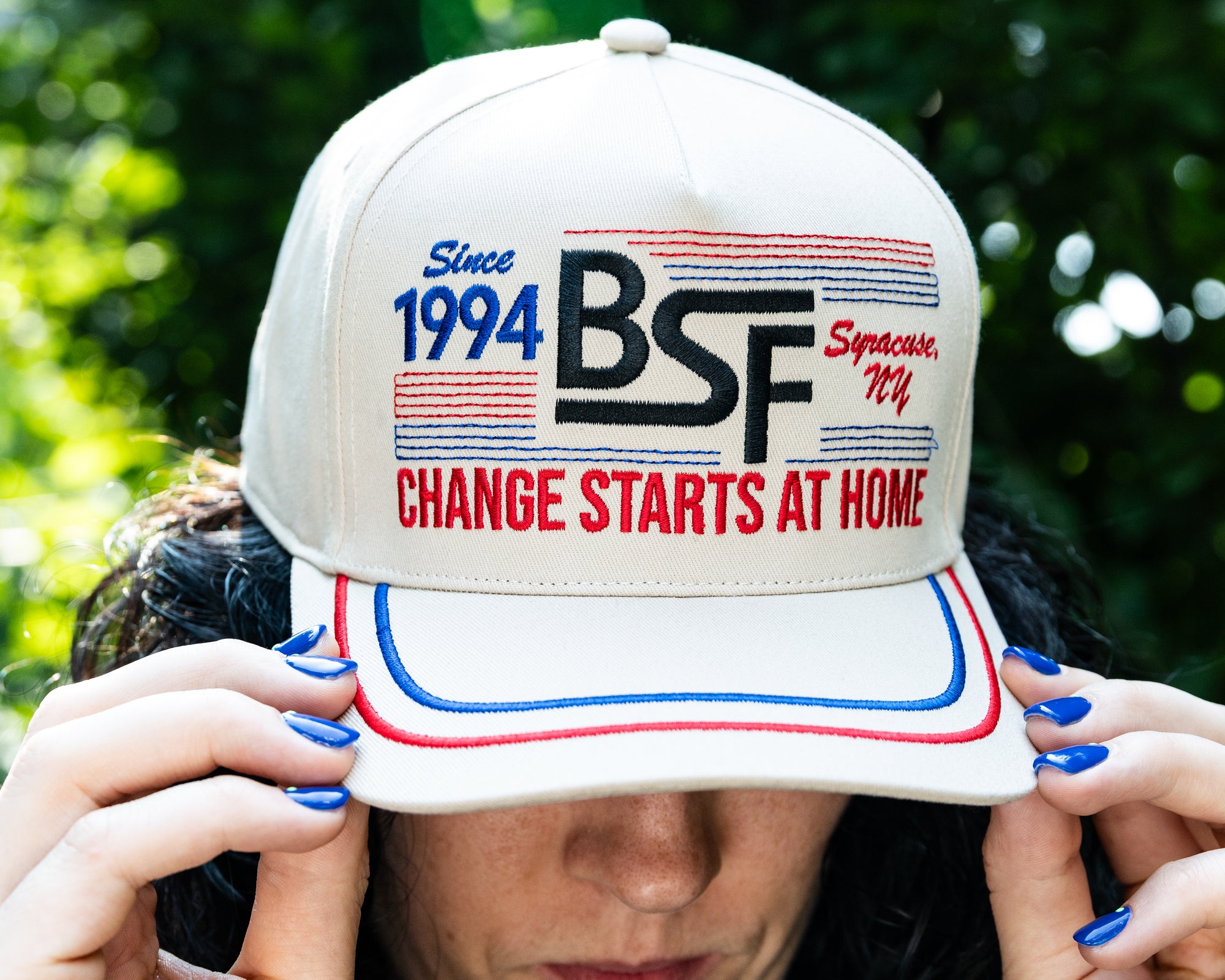 A person outdoors wearing a cream-colored baseball cap with red and blue embroidered details. The cap features the text 'Since 1994 BSF Syracuse, NY' and 'CHANGE STARTS AT HOME'.