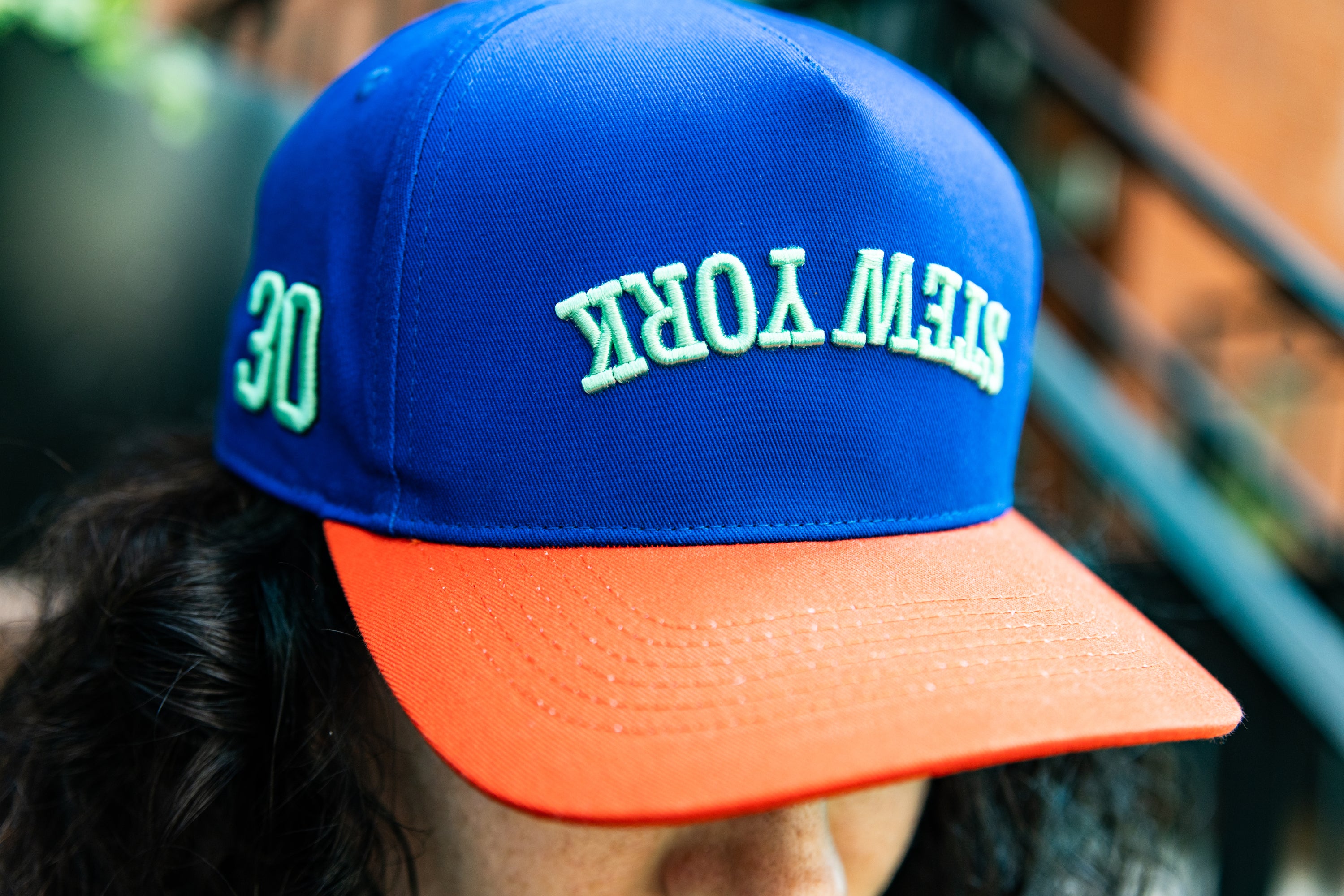 A close-up of a person wearing a royal blue baseball cap with a bright orange visor. The cap features 'STEW YORK' embroidered in light green on the front and '30' on the side.