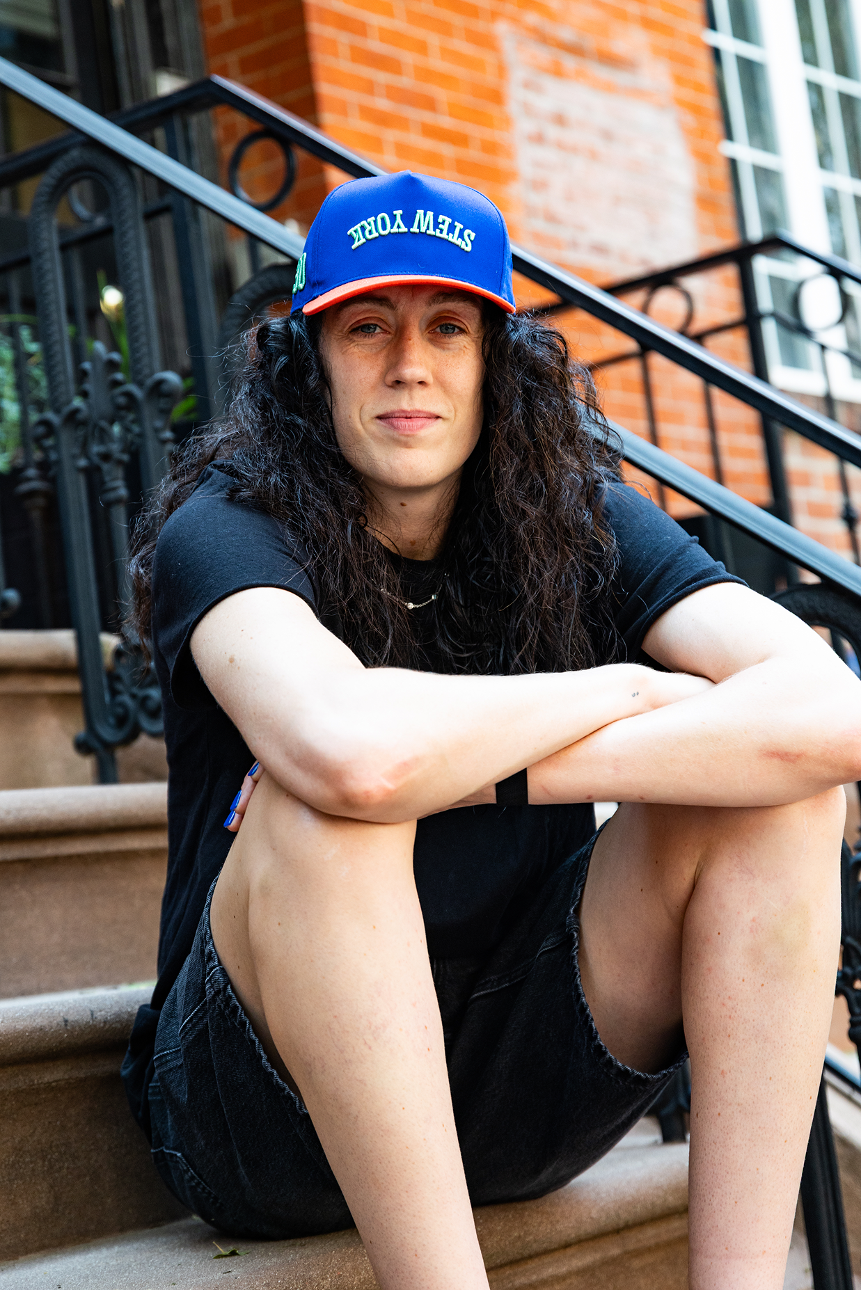 A person sitting on outdoor steps wearing a blue trucker hat, a black T-shirt, and dark shorts. The photo is taken in front of a brick building with black metal stairs.
