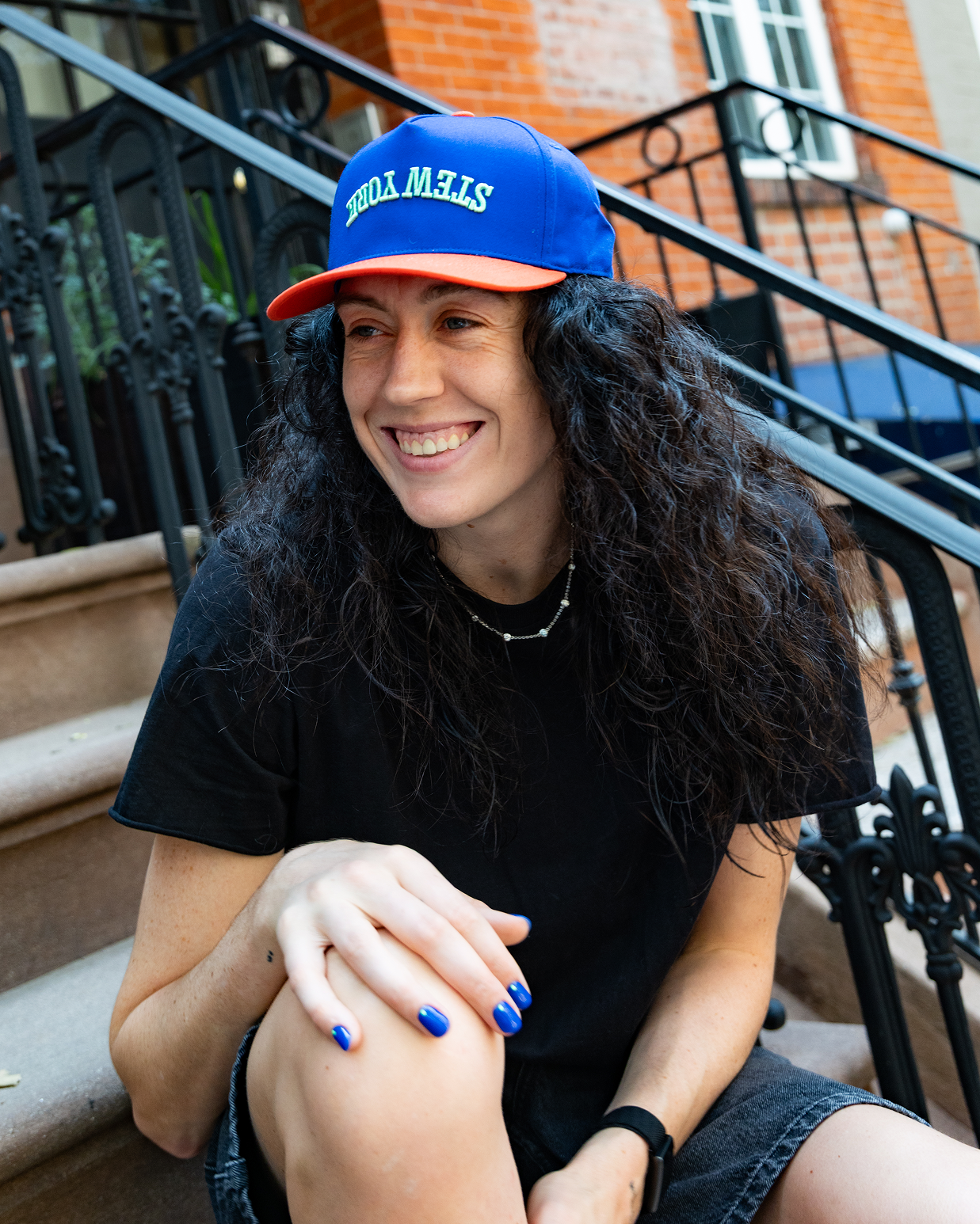 A smiling woman with long curly dark hair sits on outdoor stone steps wearing a blue and orange baseball cap a black t-shirt, and denim shorts.