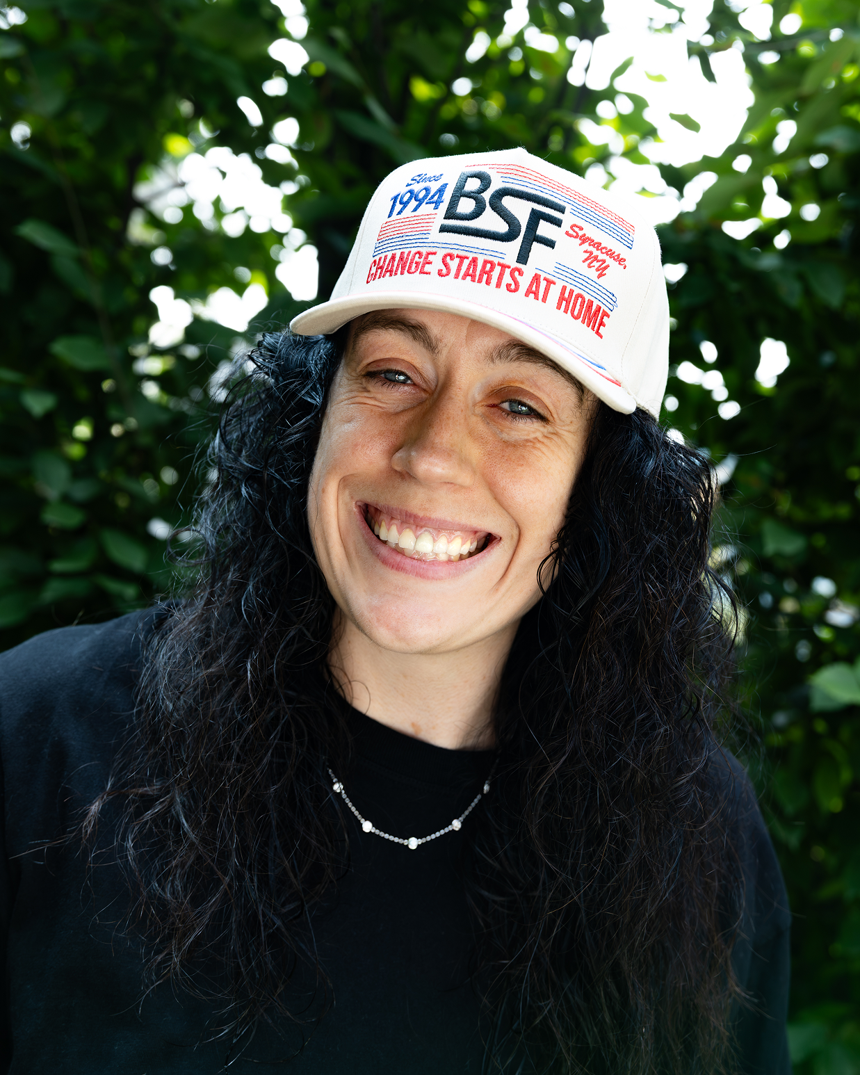 A smiling woman with long dark curly hair wearing a white baseball cap with 'BSF' and 'CHANGE STARTS AT HOME' printed on it, photographed outdoors against a blurred green leafy background.