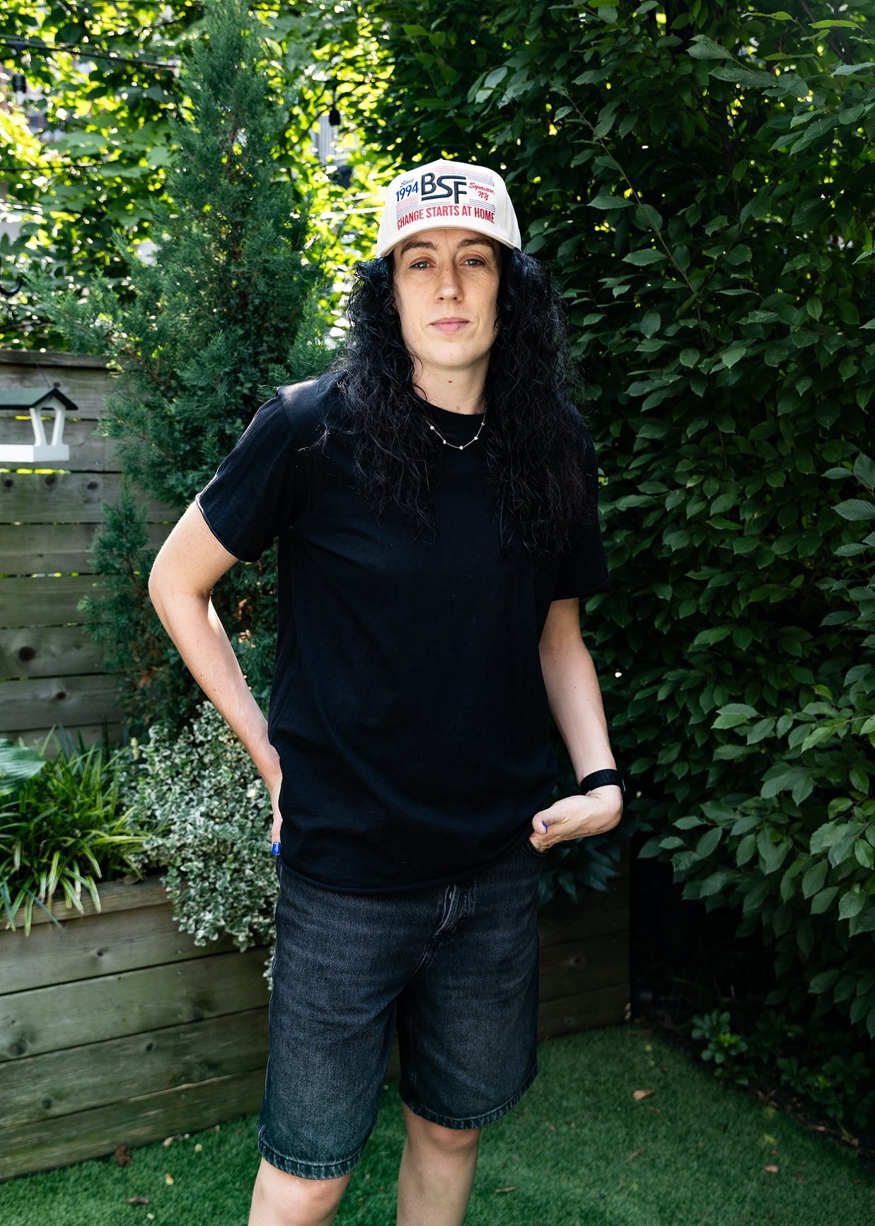 A person standing outdoors in a backyard setting, wearing a black t-shirt, dark denim shorts, a black watch, and a white baseball cap with red and blue text that reads 'THE BSF'
