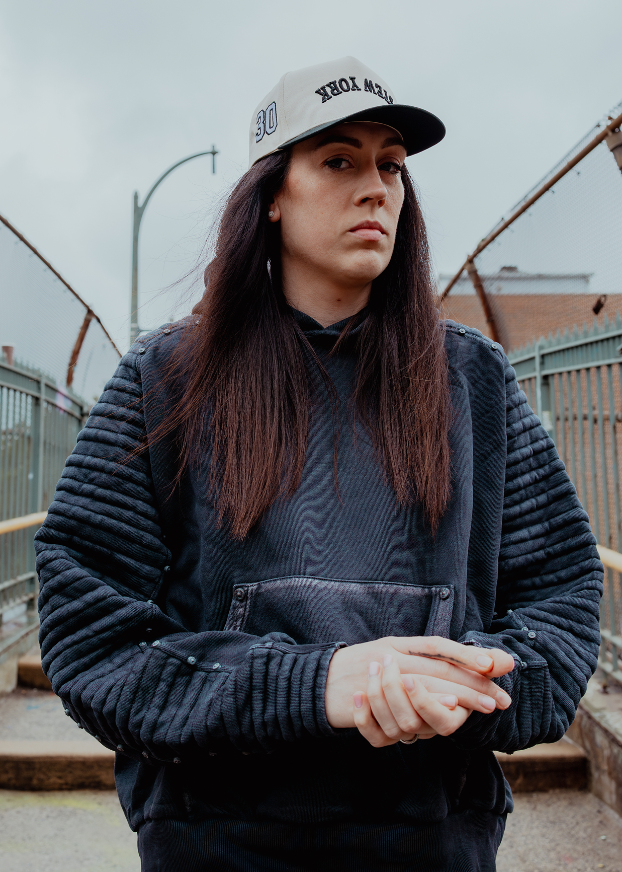 A woman wearing a dark charcoal gray quilted hoodie with a front pocket and a white trucker hat with black text, standing outdoors against a cloudy sky and metal fencing.