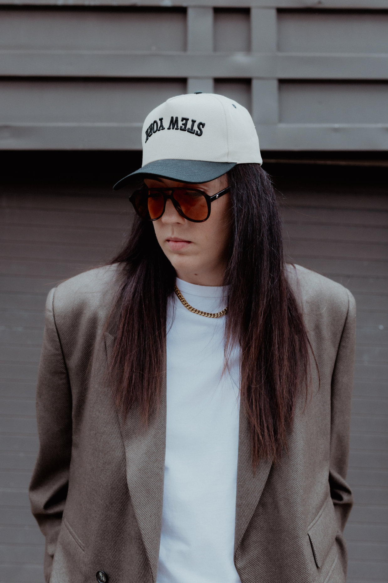 A person with long dark hair wearing a taupe blazer, white t-shirt, gold chain necklace, black sunglasses, and a white cap with black text, standing in front of a gray wooden wall.