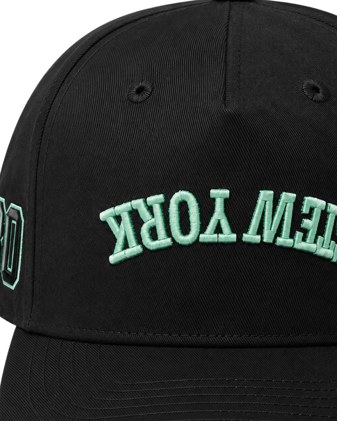 Close-up of a black baseball cap with mint green embroidered lettering that reads 'NEW YORK' upside down on the front panel.