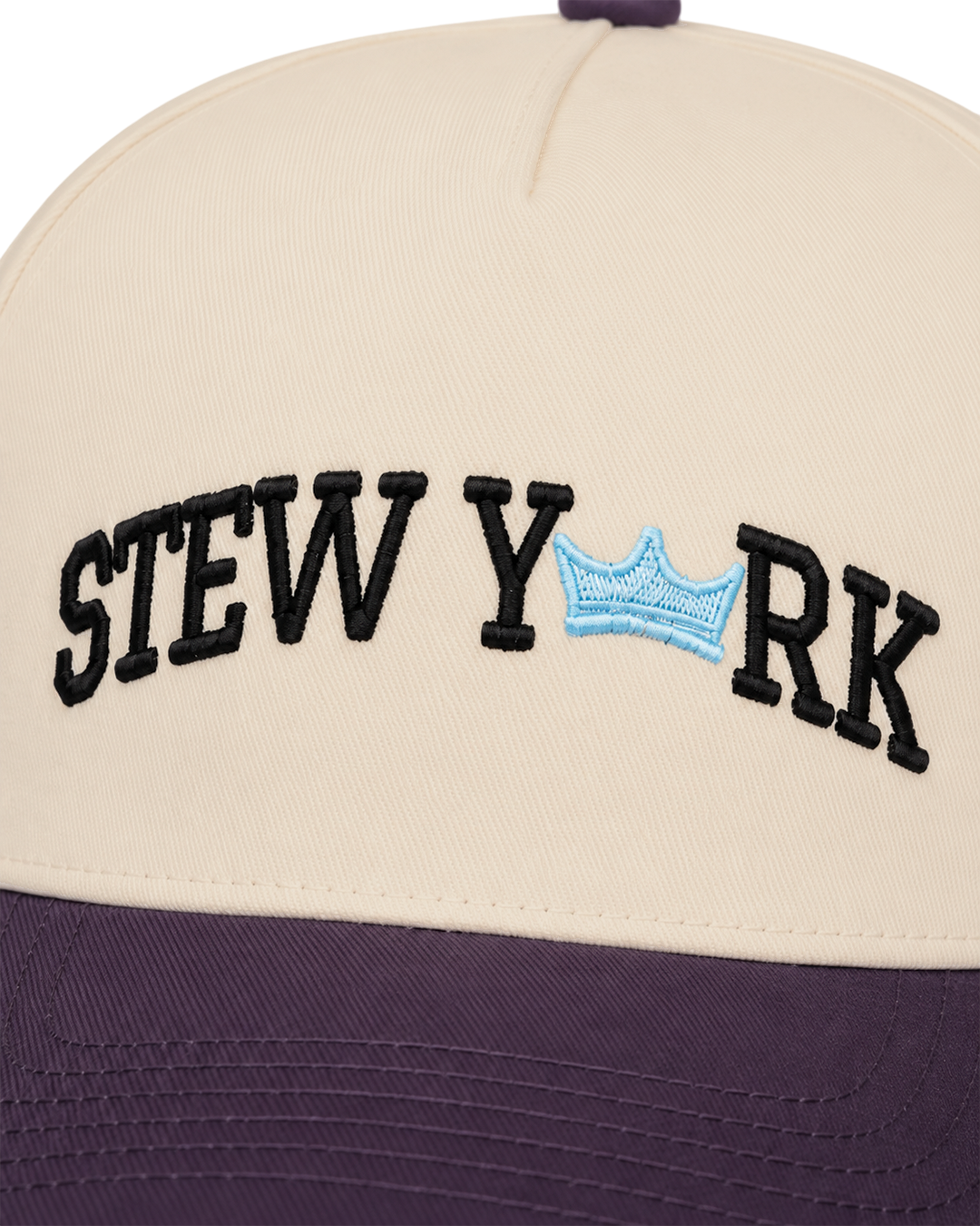 Close-up of a baseball cap with a cream crown and dark purple brim. The front has black embroidered text reading 'STEW YORK' with a small light blue crown motif between the words.