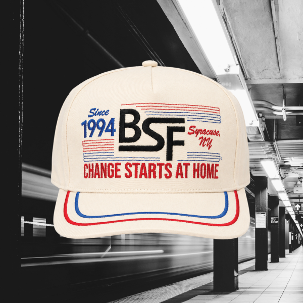 An off-white baseball cap with red and blue embroidered graphics and the text 'Since 1994 BSF Syracuse, NY' and 'CHANGE STARTS AT HOME', shown against a blurred subway station background.