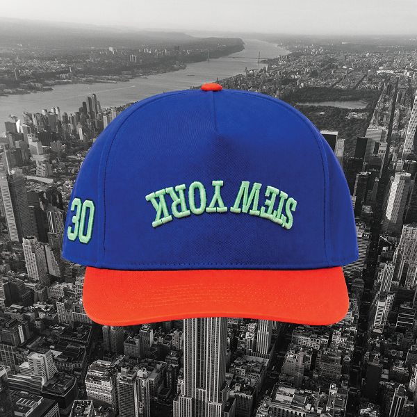 A blue baseball cap with a bright orange-red visor and green embroidered 'STEW YORK' text on the front, shown over a black-and-white city skyline background.