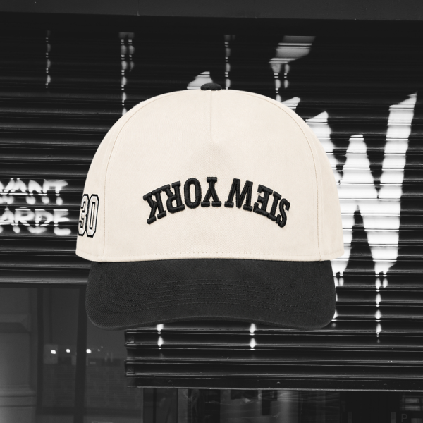 A baseball cap with a cream-colored crown and a black visor, featuring the text 'STEW YORK' in black arched embroidery on the front. The cap is shown against a dark, blurred storefront background.