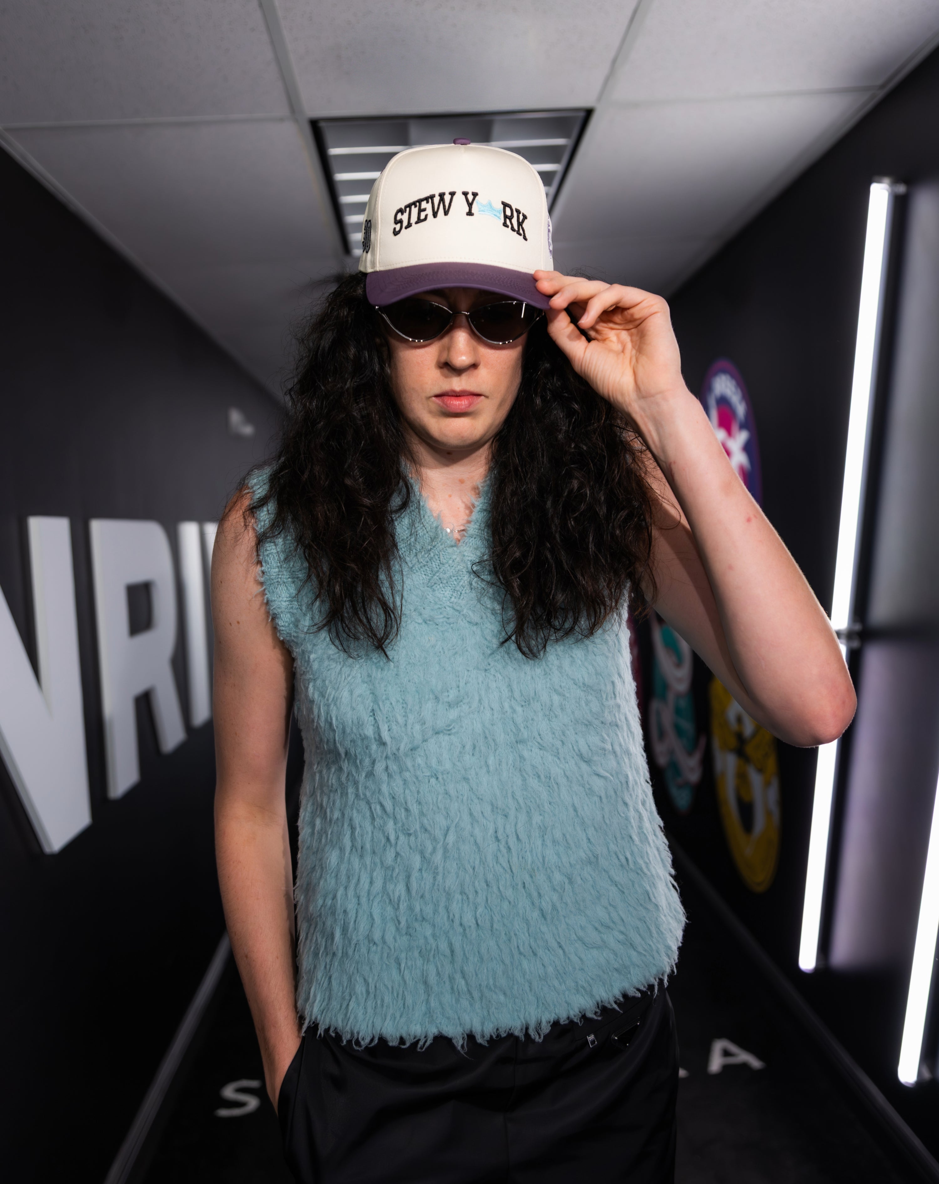 Breanna Stewart featured wearing a blue sleeveless top and white cap with 'Stew New York' text, standing indoors.
