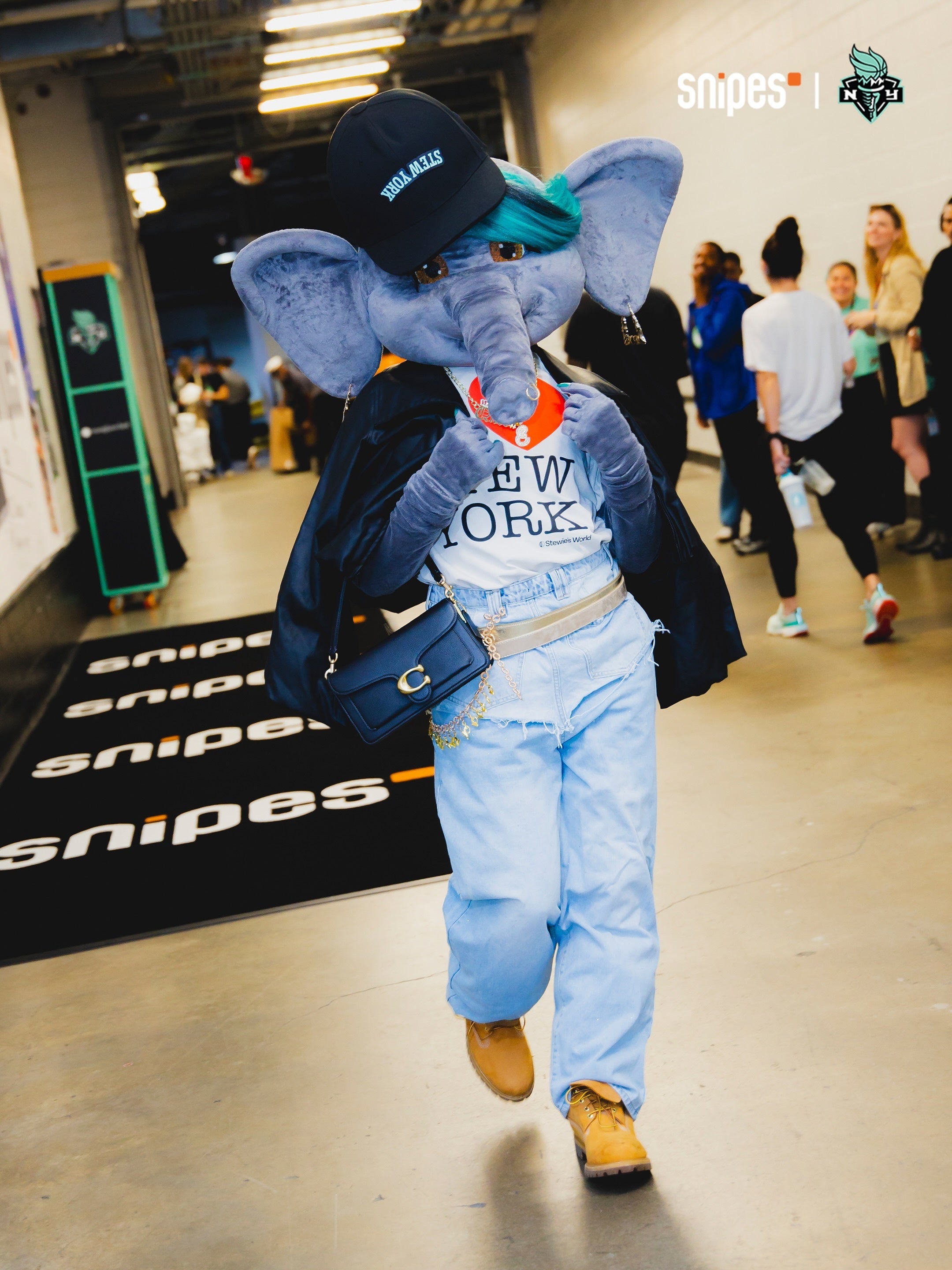A person walking indoors wearing a large gray elephant plush mask, a black cap, a white 'STEW YORK' graphic T-shirt, light blue jeans, a black jacket draped over the shoulders, and tan boots. The background shows a hallway with other people and Snipes branding on the floor and as a watermark.